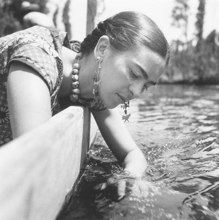 Monochrome photograph featuring a woman, upper half, leaning over a dock to place her hand  in the ripples of a river