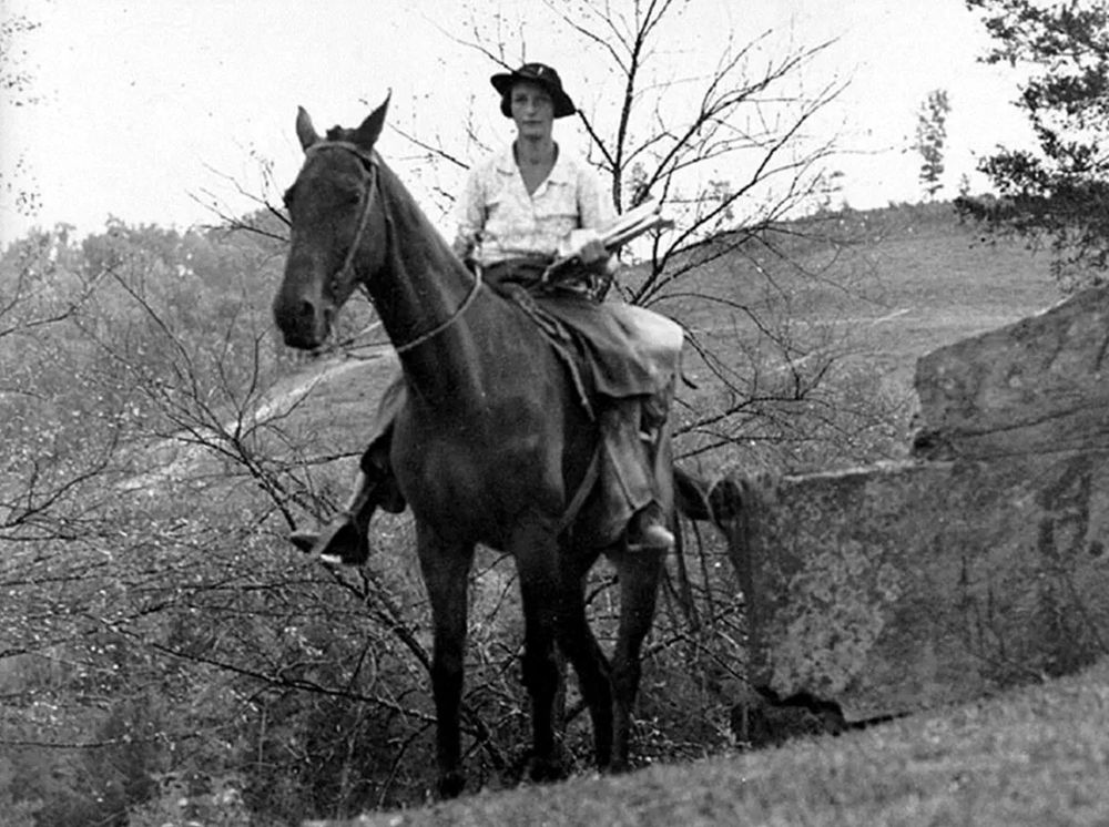 Monochrome photograph featuring a white woman with saddlebag on a black horse, facing forwards, in a rural environment 