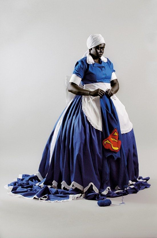 Figure of a standing Black woman dressed in a long blue dress with white apron and hat, holding  a piece of material with a superman logo, all in a white interior 