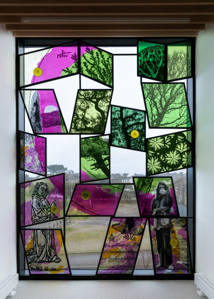 Artwork featuring a window with rectangular pieces of coloured glass with images of plants and trees in greens and pinks in a framed work with spaces between showing the real view from the window