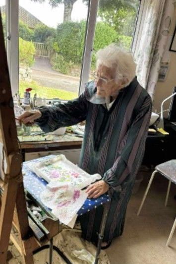 Photo of an elderly white woman facing left in an interior space, next to a window tbrough which is a green garden, in the act of painting
