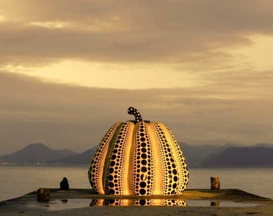 Sculpture of a yellow pumpkin with black dots on a quay next to water with hills on a distant shore behind, all in dim light