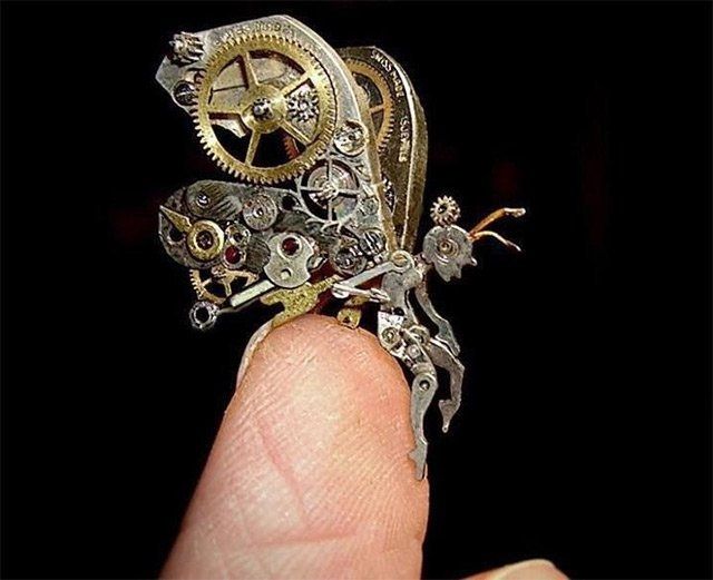 Tiny figure with large wings created with cogs and other metal small parts sitting on the end of a white real finger