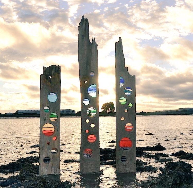 Three oart sculture created with vertically sited pieces of driftwood inlaid with circles of coloured stained glass, placed on a riverbank against a gooden sky