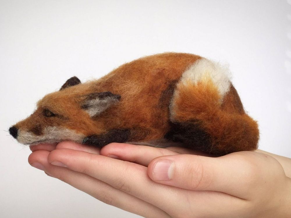 Smal furry sculpture of a fox lying with head flat facing left, resting on a real white hand in the photo 
