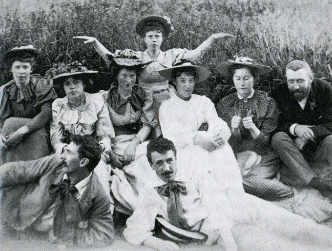 Monochrome photograph of a group of six women and three men sitting on the edge of a field with long grass dressed in Victorian/Edwardian style fashions including the women in large hats, most are facing forwards, a woman at the back holds her arms outstretched 