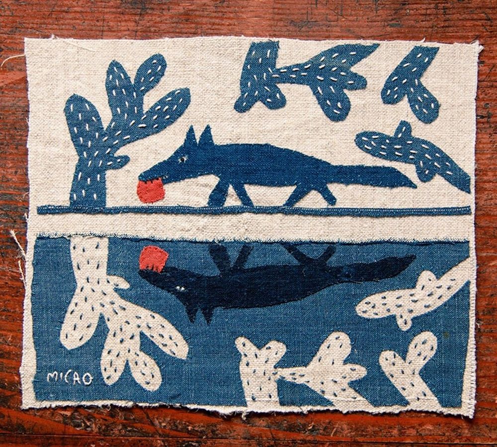 Textile artwork with patched naterial and visible stitching featuring a stylised blue fox moving to the left with an apple in its mouth, among trees, all perfectly reflected in a pond below