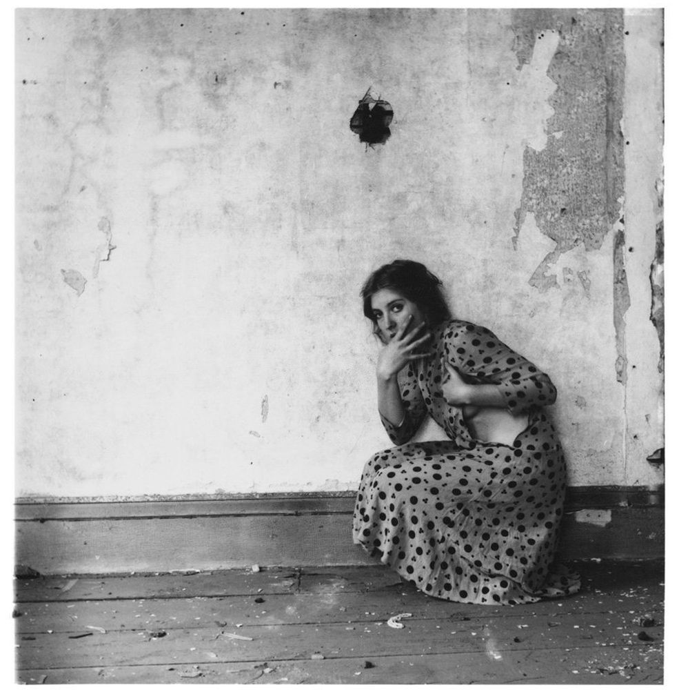 Monochrome photograph featuring a crouching white woman in a spotty dress next to an intetior wall with torn wallpaper
