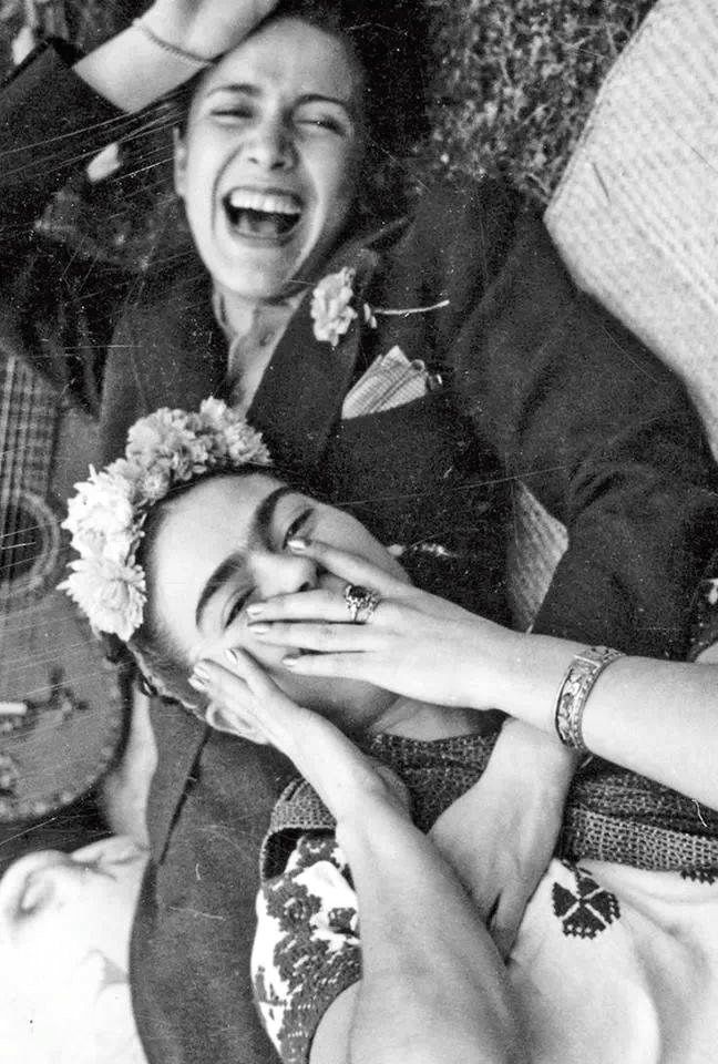 Monochrome photograph of the upper halves of two women lying on the ground entwined together and laughing, one subject wears a jacket and has one hand on her head and the other across the chest of the other figure, the other figure wears a white patterned top and has flowers in her hair, she holds her hands over her mouth