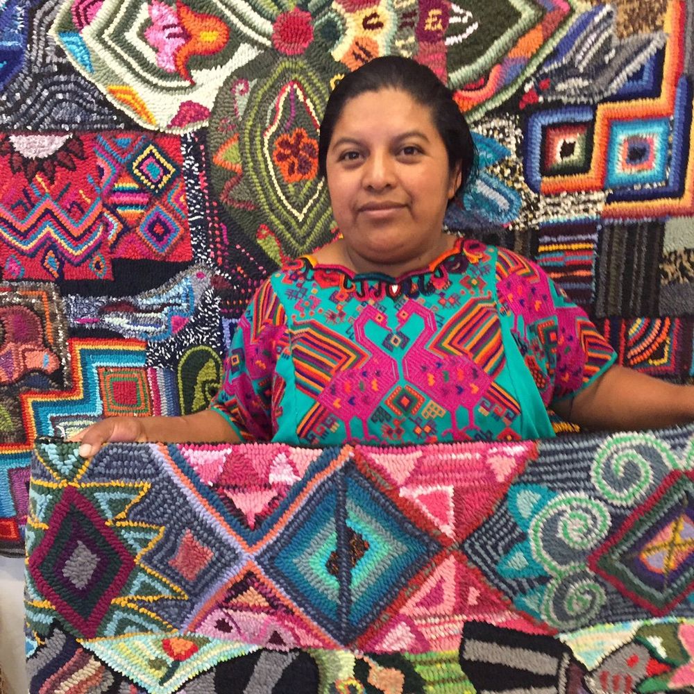 Photo of a woman with dark hair holding a patterned colourful wpoven textile with more woven textiles behind her