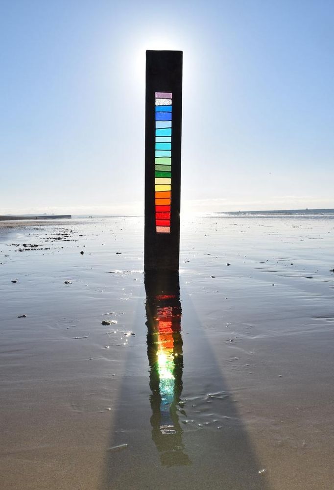 Photo of a slim rectangular wooden sculpture with central stained glass in rainbow colours the artwork is on a beach with wet sand and under a blue sky