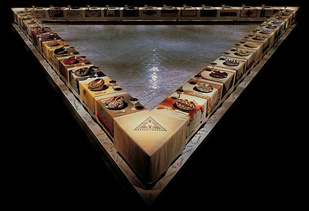 Photo of a large-scale installation in a gallery, the work features a huge triangular shaped table set with individually created ceramic plates and embroidered cloth name places with the names of notable women from history, the whole work is under light in a dark room