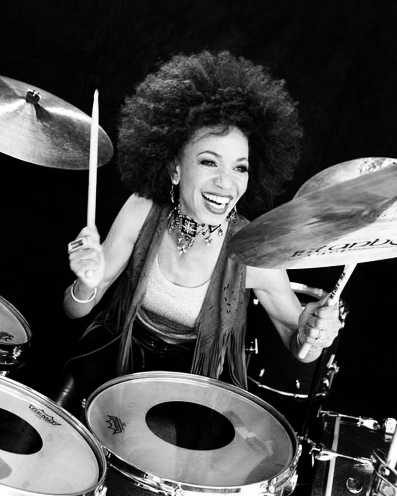 Monochrome photograph featuring a close-up of a Black woman sitting at a drumkit in the act if playing drums  wearing a vest top and waistcoat and smiling