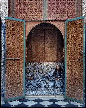 Photograph featuring a woman lying on a bed within an arched space with large open doors decorated with geometric tile designs 