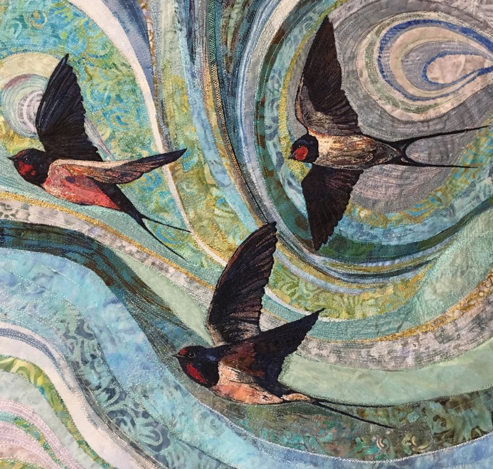 Textile artist featuring three small black and white birds with long forked tails in flight, flying right against a swirling blue green sky