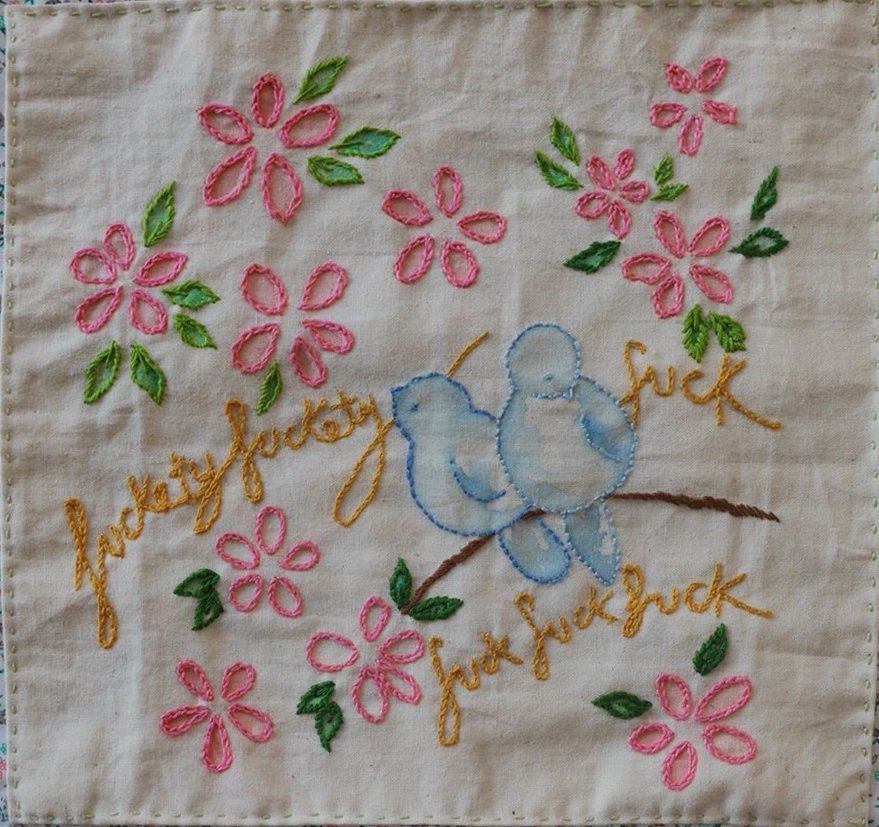 Embroider of two small blue birds perched among pink flowers with embroidered text in gold thread stating the title