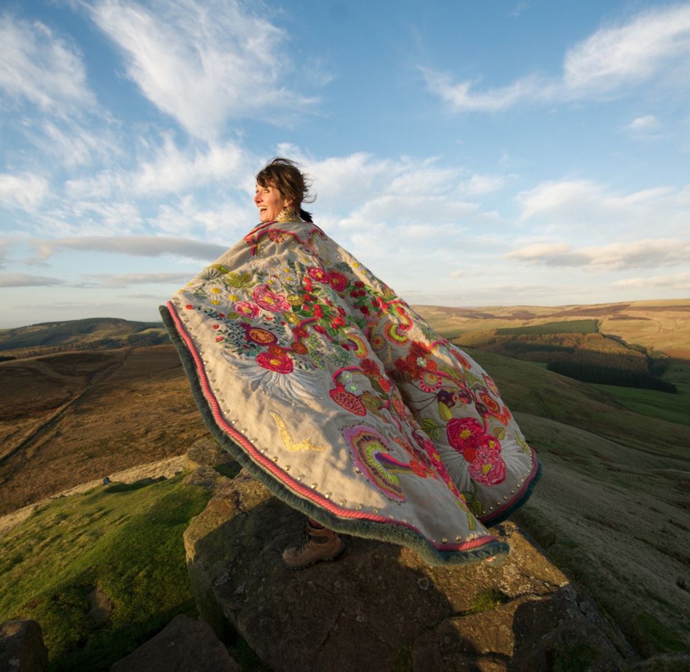 Photo of a smiling white woman on a windswept high moorland landscape in a large textile art cloak with patterns and motifs which lifts slightly in the wind