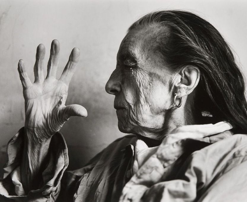 Monochrome photograph of an older white woman, head and shoulders, facing left with right hand held in front of her face with open palm, the subject has eyes closed and long dark hair and wears a ruffled shirt, all against a plain background 