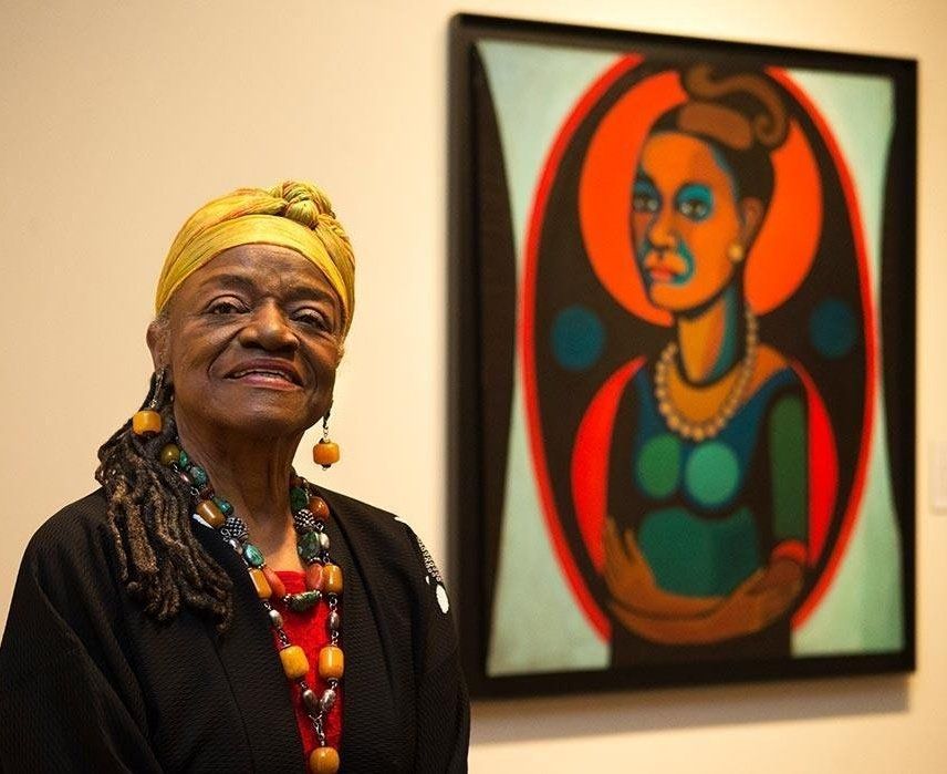 Photograph featuring the upper half of a standing Black woman, to the left, looking outwards with head raised proudly, to the right is her self-portrait, upoer half looking left,  on a cream wall 