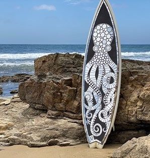 Photo featuring a surfboard, with octopus design in white on black, propped upright against rocks on a beach by the sea