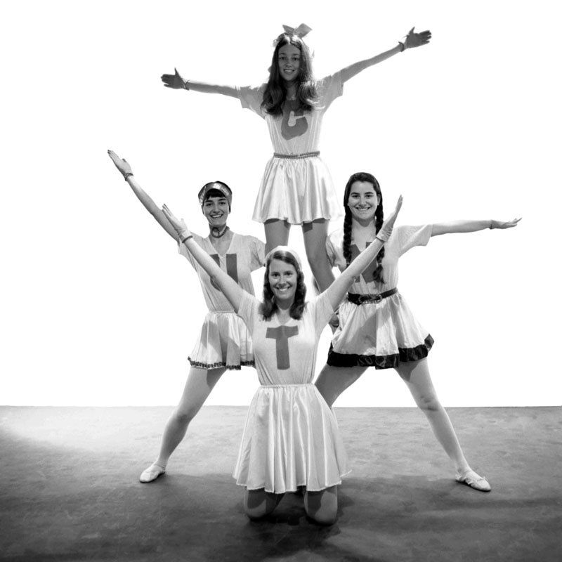 Monochrome photograph featuring four white women in cheerleader style outfits with a letter on the front of each, preforming an acrobatic display
