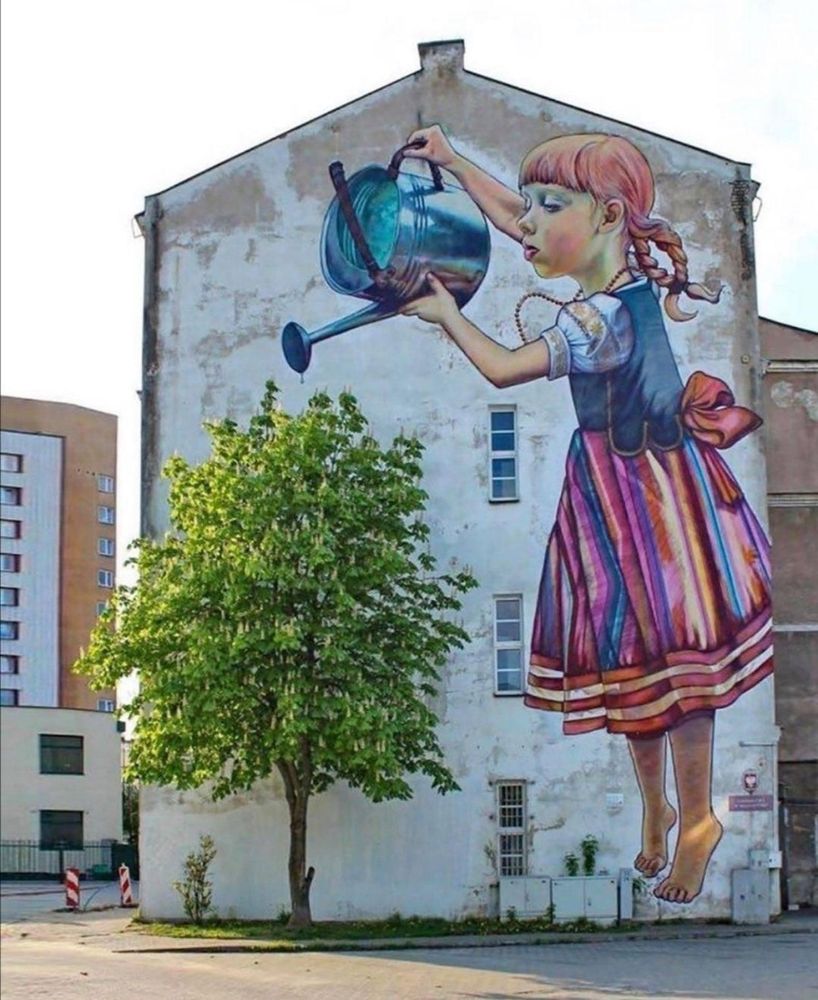 Photo featuring the side of a large building with a huge mural of a white girl in a dress holding a watering can and tipping it over a real tree which grows by the side of the building on an urban street 