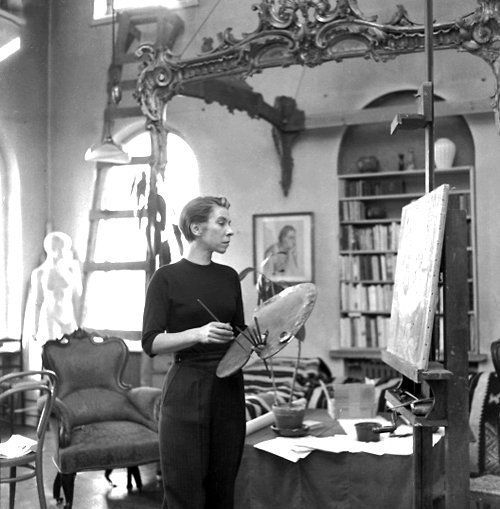 Monochrome photograph featuring a standing white woman in an interior with art equipment and bookshelf, the subject wears dark top and trousers, faces right, holding brushes about to paint on a canvas