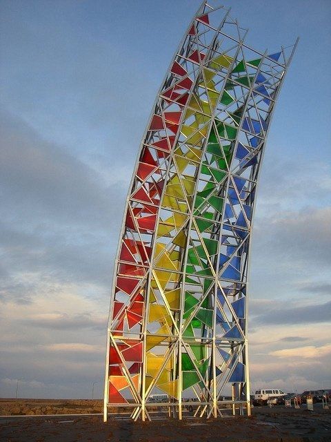 Photo of a large scale sculpture sited outdoors, on a flat landscape under a blue cloudy sk   in the shape of a section of a rainbow created with steel arches and stained glass panels
