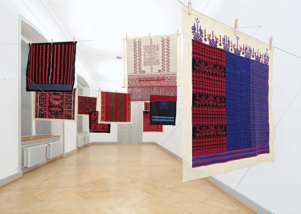 Photo of a series of square colourful textile pieces hanging on lines at angles in a narrow white gallery room  