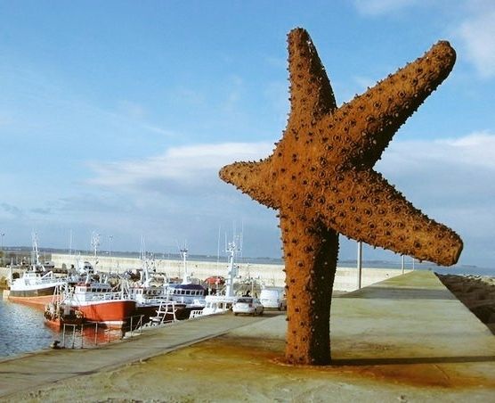 Standing starfish sculpture on the side of a quay next to the sea and harbour with small boats