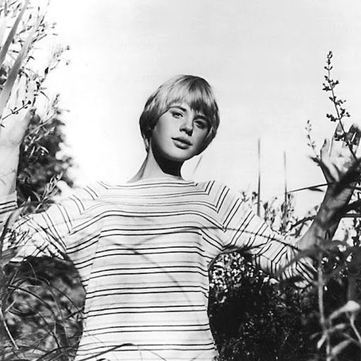 Monochrome photograph featuring a young white woman outdoors, upper half, facing forwards among foliage, with arms held up to her sides, she has short blond hair and wears a light coloured striped t-shirt 