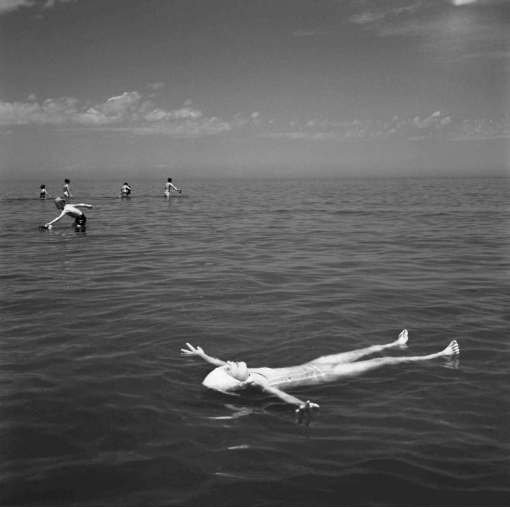 Monochrome photograph featuring a white woman in a bathing costume floating on her back on the surface of a lake or sea