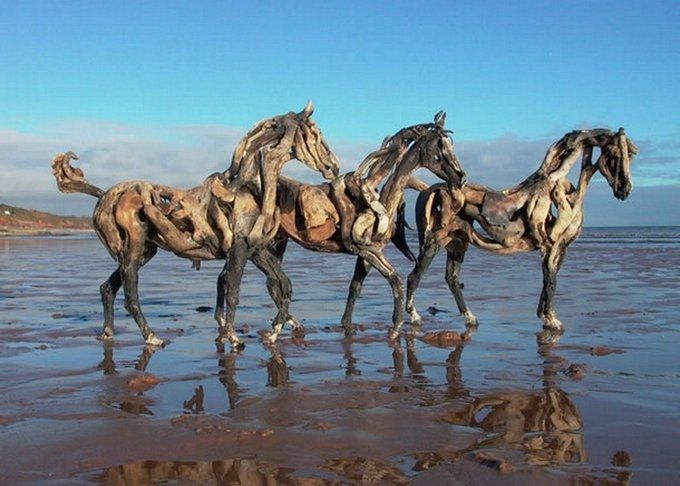 Photo featuring three horse sculptures grouped together on the wet sand of a beach under a blue sky, each horse is created with pieces of drift wood and stands facing right