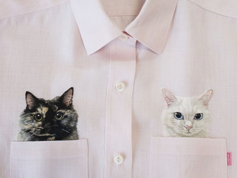 Photo of two realistic looking embroidered cats heads,  one blackish, one white, proving out the top of pockets on a white shirt