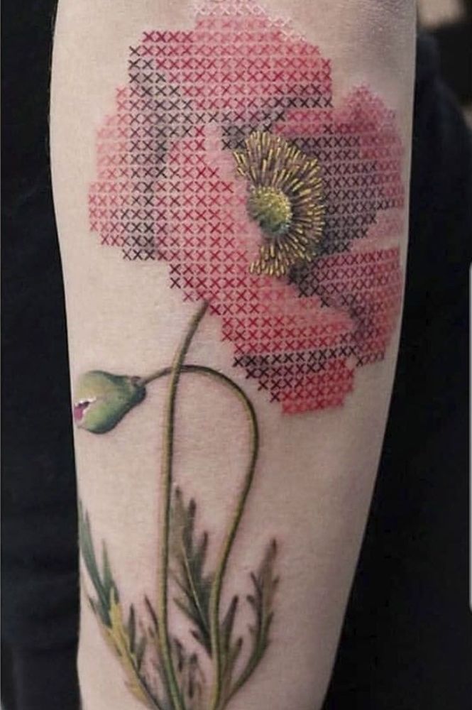 Photo of a tattoo of a red poppy with stem and leaves, on a white arm, the tattoo has many littke crosses on the petals imitating sewn cross stitches