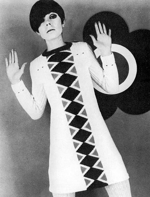 Monochrome photograph featuring a standing white woman with hand up and dark bob hairstyle wearing a short white dress with central vertical diamond motif