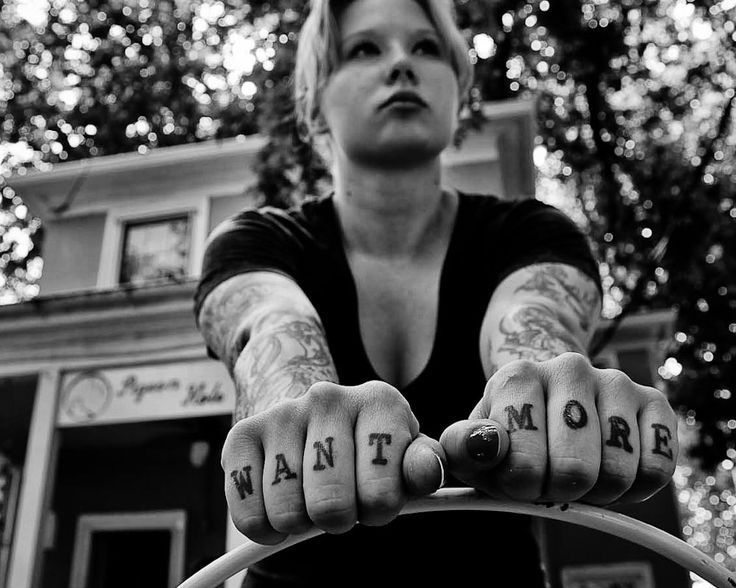 Monochrome photograph featuring a standing white young woman with the words 'want' and 'more' tattooed on her knuckles