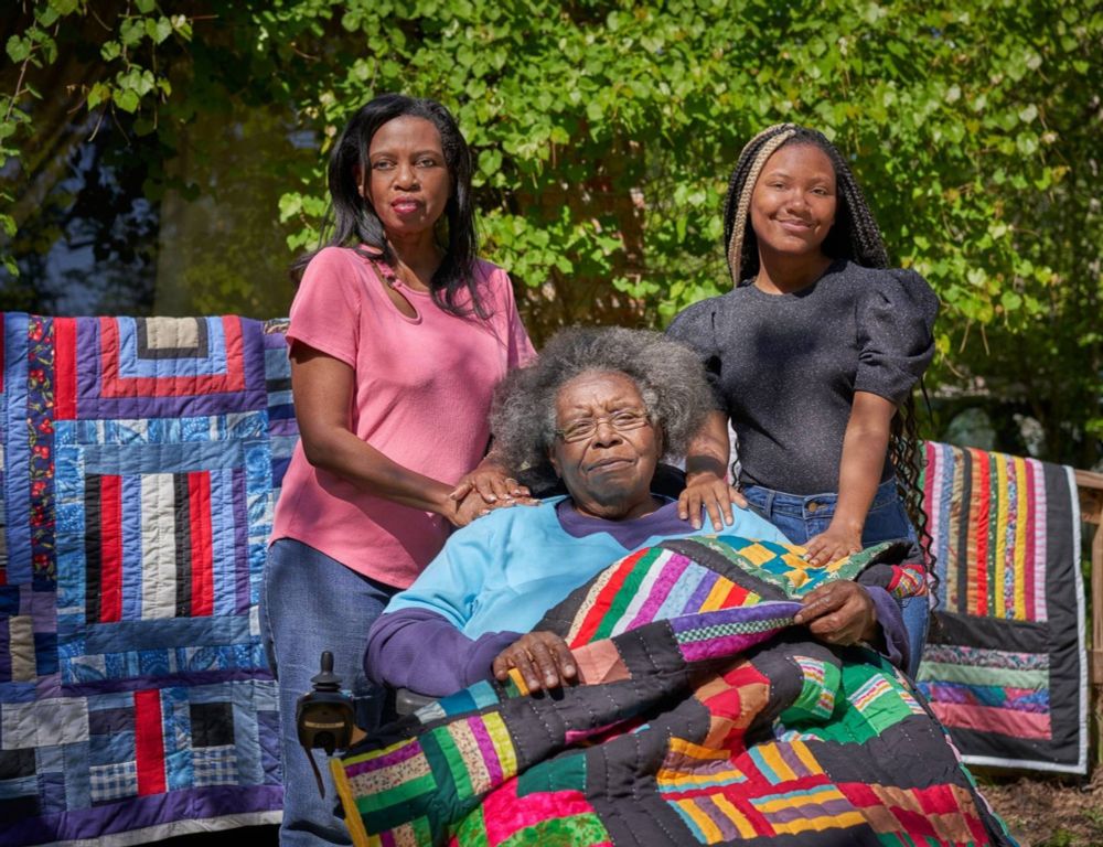 Photo featuring three Black women in an outdoor space by trees, two subjects stand either side of the third who sits in the center with a quilt on her knee, to the sides are more colourful quilts