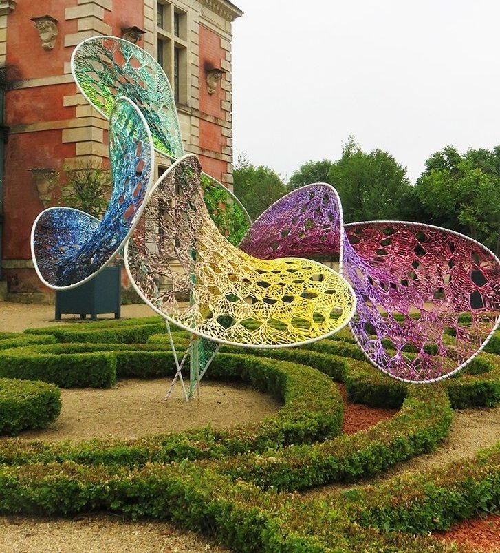 Photo of wire and woven sculpture with four bent circular parts sited in a formal garden above with clipped hedges and a grand red stone building behind
