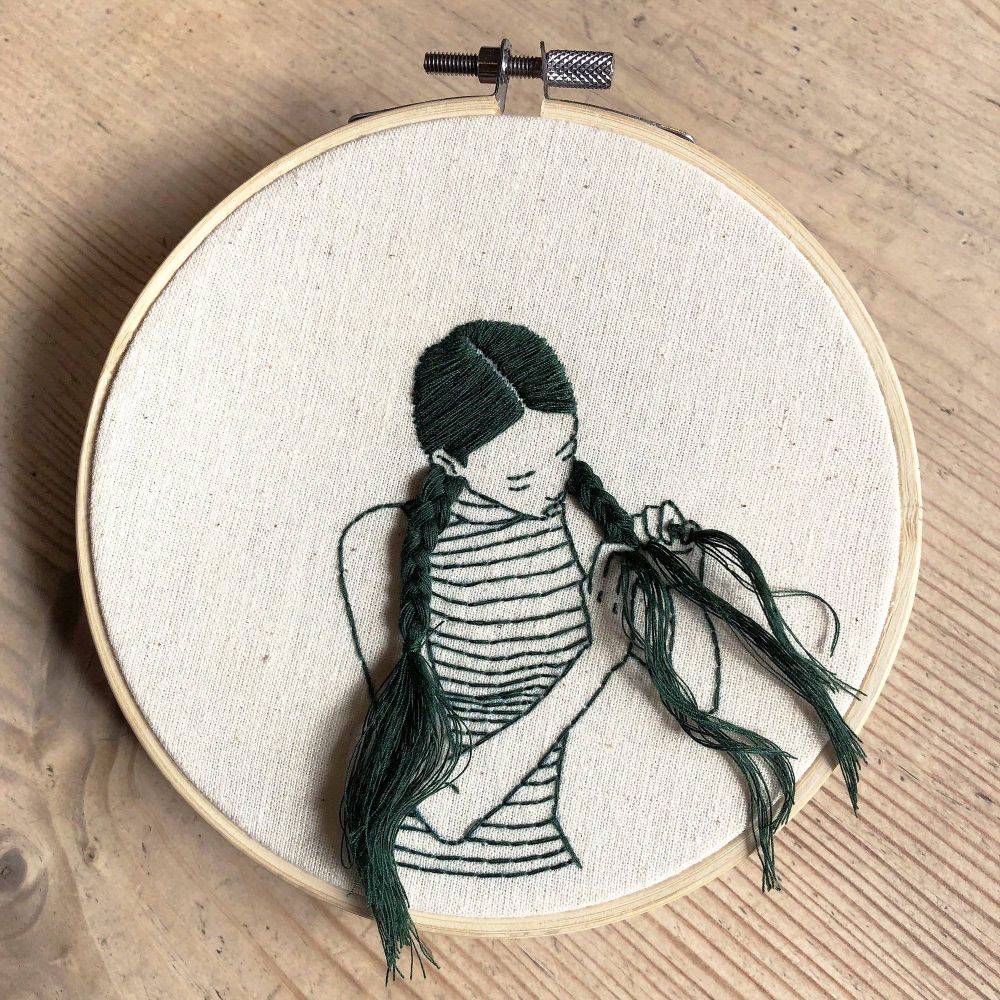 Photograph featuring an embroidery hoop on a pale wooden surface with an image of a figure in black thread sewn on tbe cream material the figure  a young woman  is shown braiding her long hair with thread that produdesqq from the image and wears a striped top
