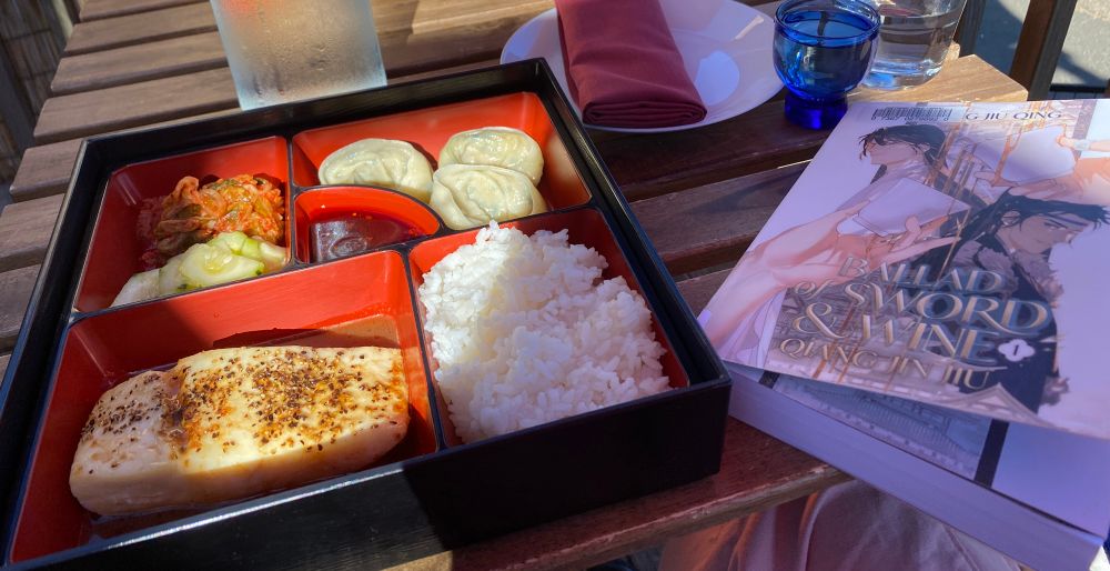 Photo of a wooden table with a copy of “Ballad of Sword and Wine Volume 1” by Tang Jiu Qing on top of it, next to a scrumptious bento box with assorted side dishes, rice, and fish.