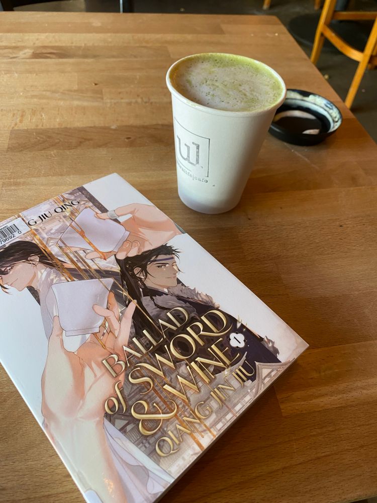 The first volume of the novel “Ballad of Sword and Wine”  by Tang Jiu Qing on a wooden tabletop next to a scrumptious little matcha latte.