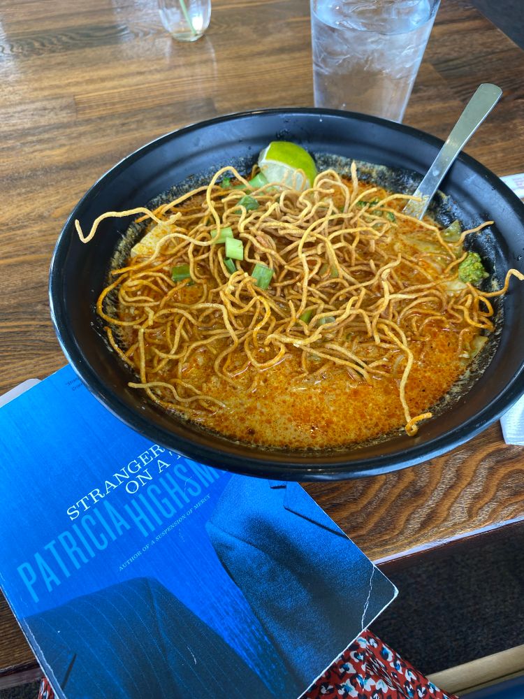 Strangers on a Train by Patricia Highsmith on a wooden tabletop next to a scrumptious looking bowl of khao soi noodle soup.