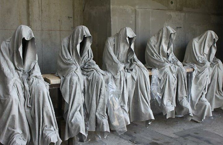 A spooky sculpture installation showing five robes that seem to be filled with invisible forms sitting on a bench