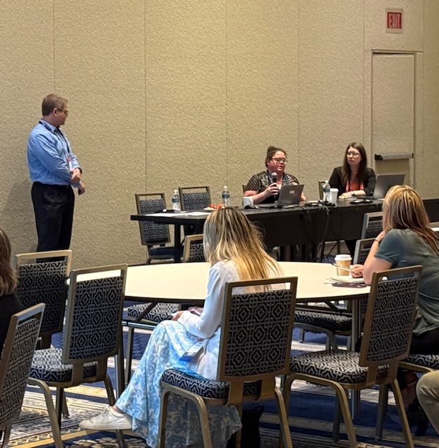 FAVOR's Lisa Girard speaks from a table at the front of the training room as CHDI's Jason Lang and Kellie Randall look on
