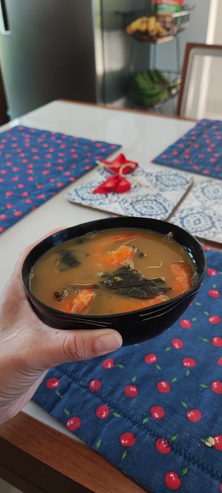 A imagem mostra uma cuia contendo tacacá, um prato típico da região Norte do Brasil, feito com caldo de tucupi, camarões, jambu e goma de mandioca. Estou segurando a cuia sobre uma mesa decorada com um tecido azul estampado com cerejas. Ao fundo, é possível ver uma fruteira com bananas e parte de uma cozinha.


