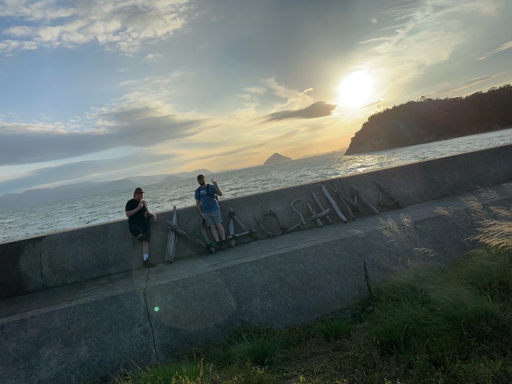 A photograph showing a wave barrier dividing the concrete foreground from the ocean beyond. The photo is on a slant. In the top half, the sun is beginning to set over the ocean. A small island is visible in the far distance, and an outcropping of the island is more prominent closer to shore. The sky is overcast but clouds are thinning. 

In the bottom half of the photo, land is visible showing a smooth concrete barrier that is waist high. Driftwood spells out Naoshima. Standing before the N is one individual showing a piece sign in a black shirt. After the N is a second person in a blue shirt. 