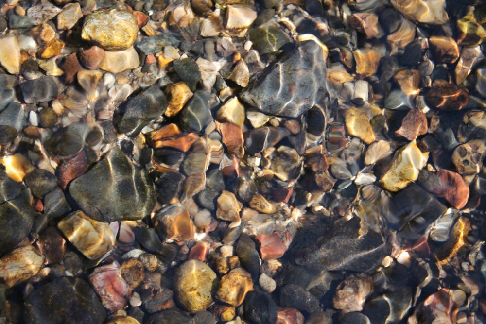 A photo through a shallow stream of brightly colored rocks with light refracting through the water onto them.