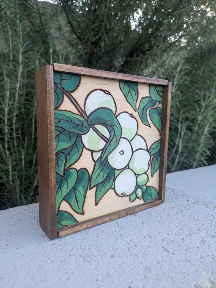 An angled shot of a wood burning of a cluster of berries and leaves on a wood panel, colored using watercolor. There's a rosemary bush in the background. 