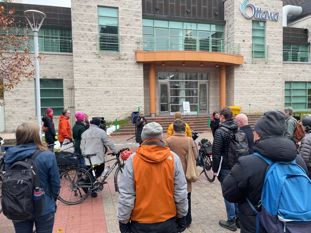 Florence from Bike Ottawa is seen in the background speaking at the rally in front of City Hall. In the foreground, you can see more people and their bikes. 